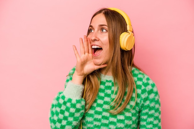 Young caucasian woman listening to music isolated on pink background shouting and holding palm near opened mouth