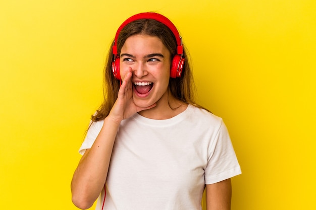 Young caucasian woman listening to music isolated on yellow background shouting and holding palm near opened mouth