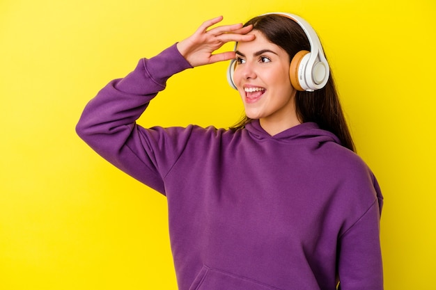 Young caucasian woman listening to music with headphones isolated on pink wall looking far away keeping hand on forehead.