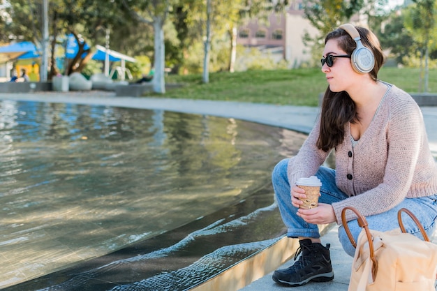 公園でコーヒーを飲みながらヘッドフォンで音楽を聴いている若い白人女性