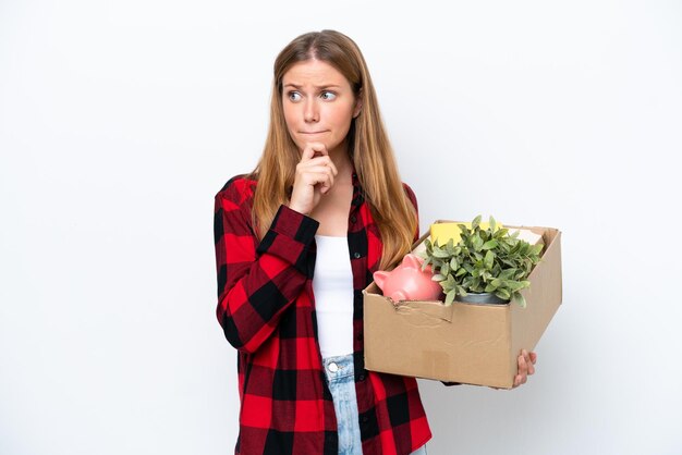 Young caucasian woman making a move while picking up a box full of things isolated on white background having doubts and thinking