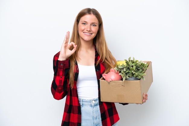 Young caucasian woman making a move while picking up a box full of things isolated on white background showing ok sign with fingers