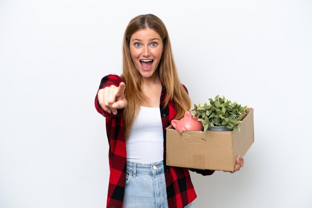 Young caucasian woman making a move while picking up a box full of things isolated on white background surprised and pointing front