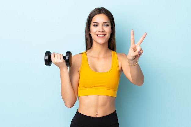 Young caucasian woman making weightlifting on blue smiling and showing victory sign