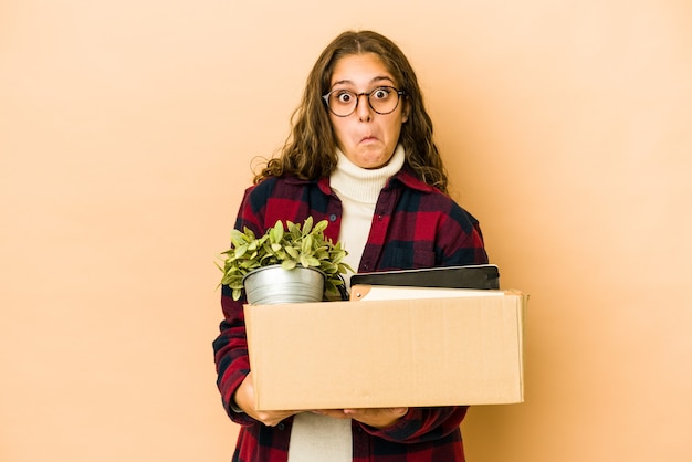 Young caucasian woman moving holding a box isolated shrugs shoulders and open eyes confused.