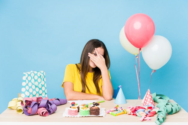 Young caucasian woman organizing a birthday blink at the camera through fingers, embarrassed covering face.