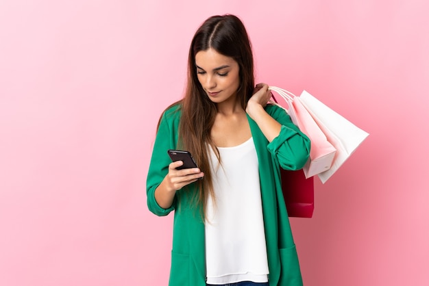 Young caucasian woman on pink holding shopping bags and writing a message with her cell phone to a friend