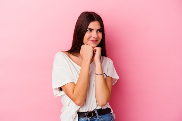 Young caucasian woman on pink keeps hands under chin, is looking happily aside.