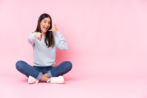 Young caucasian woman on pink wall making phone gesture and pointing front