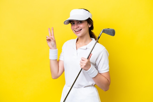 Молодая кавказская женщина, играющая в гольф на желтом фоне, улыбается и показывает знак победы