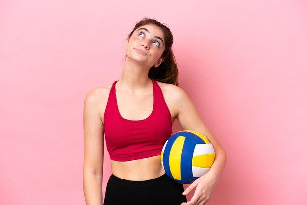 Young caucasian woman playing volleyball isolated on pink background and looking up