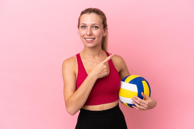 製品を提示する側を指しているピンクの背景に分離されたバレーボールをしている若い白人女性