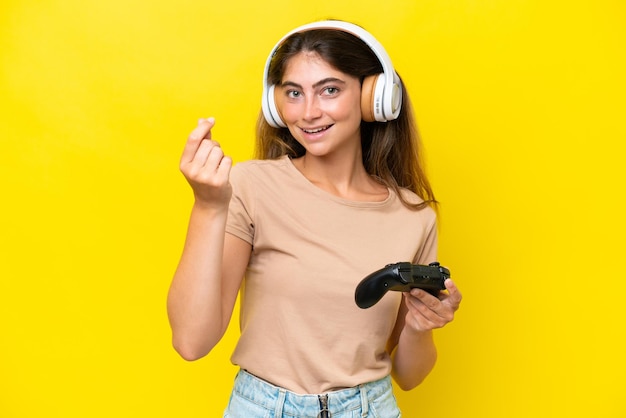 Young caucasian woman playing with a video game controller isolated on yellow background making money gesture