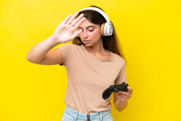 Young caucasian woman playing with a video game controller isolated on yellow background making stop gesture and disappointed