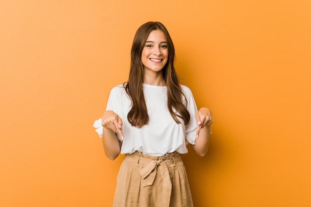 Young caucasian woman points down with fingers, positive feeling.