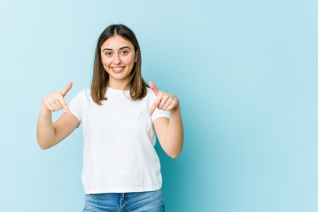 Young caucasian woman points down with fingers, positive feeling.