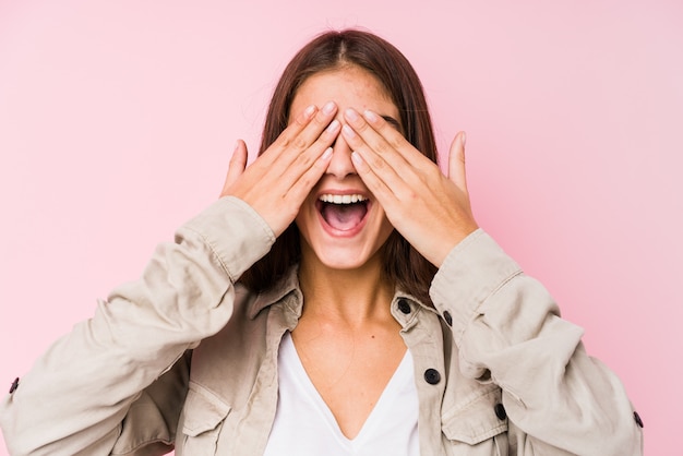 Young caucasian woman posing in a pink wall covers eyes with hands, smiles broadly waiting for a surprise.