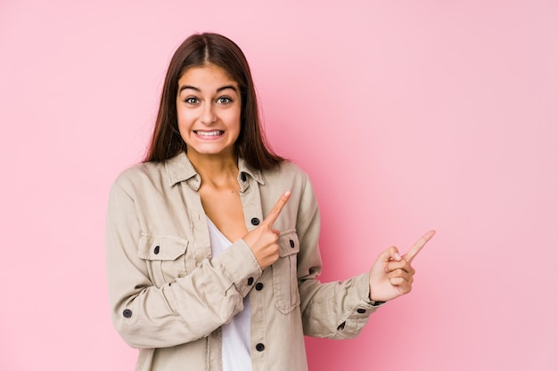 Young caucasian woman posing in a pink wall shocked pointing with index fingers to a copy space.