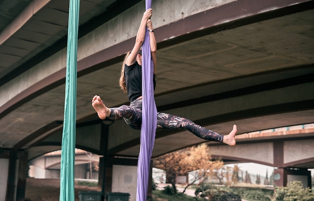 Young caucasian woman practicing aerial silks performance hanging on a bridge in a park city, surrounded by nature.