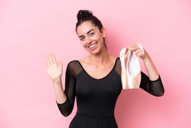 Young caucasian woman practicing ballet isolated on pink background saluting with hand with happy expression