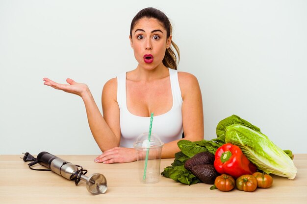 Young caucasian woman preparing a healthy smoothie with vegetables impressed holding copy space on palm.