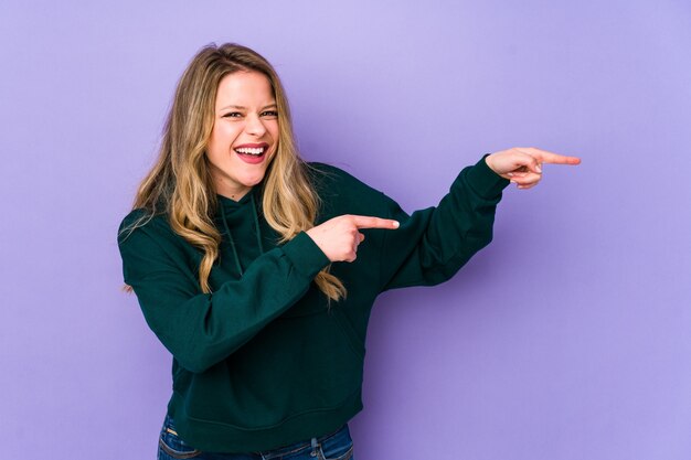 Young caucasian woman on purple excited pointing with forefingers away.