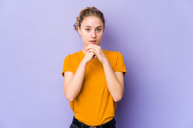 Young caucasian woman on purple praying for luck, amazed and opening mouth looking to front.