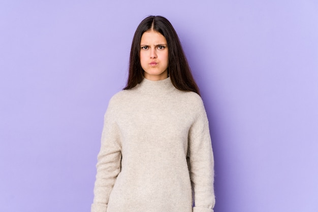 Young caucasian woman on purple wall blows cheeks, has tired expression.