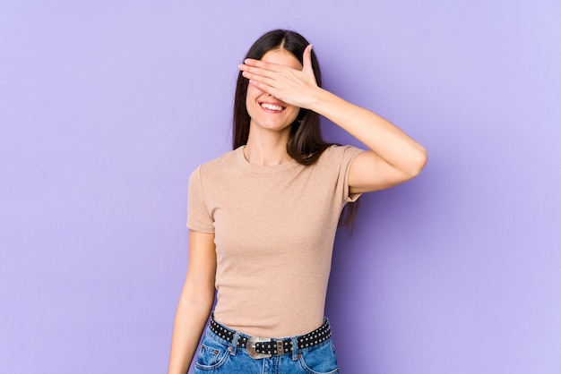 Young caucasian woman on purple wall covers eyes with hands, smiles broadly waiting for a surprise.