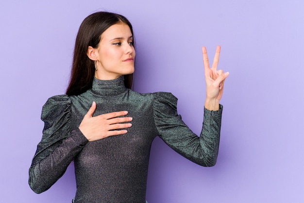 Young caucasian woman on purple wall doing victory sign, putting hand on chest.