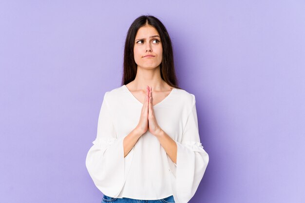 Young caucasian woman on purple wall holding hands in pray