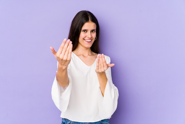 Young caucasian woman on purple wall pointing with finger at you as if inviting come closer.