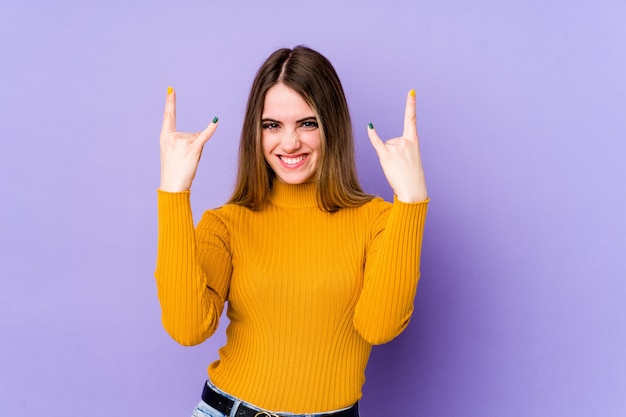 Young caucasian woman on purple wall showing a horns gesture