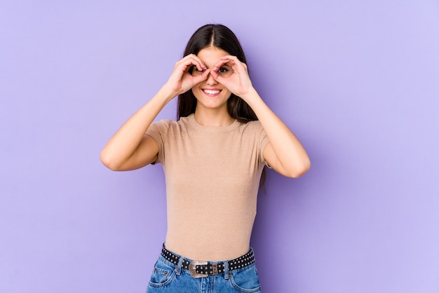 Young caucasian woman on purple wall showing okay sign over eyes