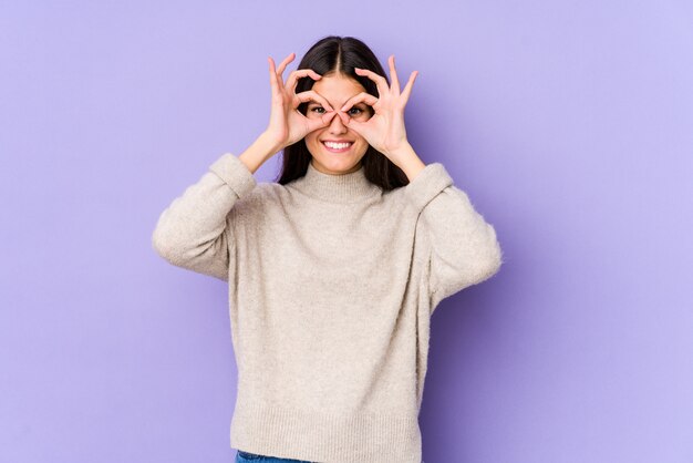 Young caucasian woman on purple wall showing okay sign over eyes