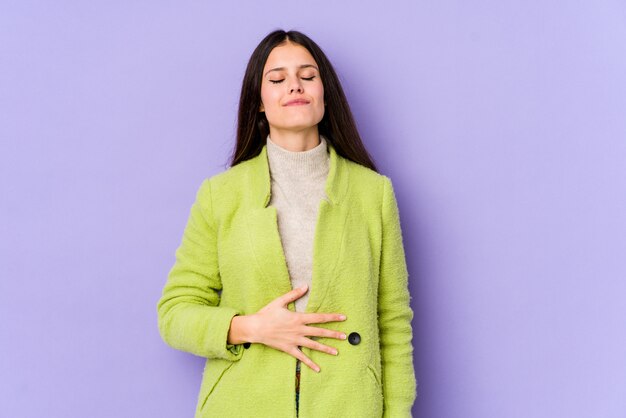 Young caucasian woman on purple wall touches tummy, smiles gently