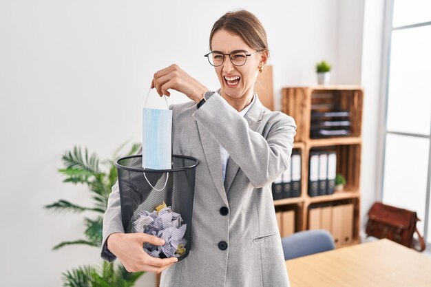 Young caucasian woman putting safety mask in the bin at the office angry and mad screaming frustrated and furious, shouting with anger. rage and aggressive concept