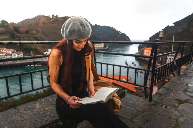 Young caucasian woman reading a book from the a viewpoint next to the sea at Pasaia; Basque Country