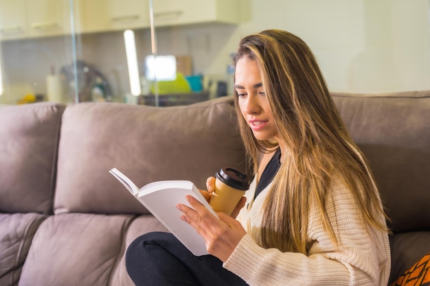 Giovane donna caucasica che legge un libro sul divano nel suo soggiorno