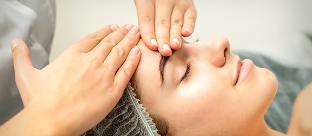 Young caucasian woman receiving facial massage by beautician's hands in spa medical salon