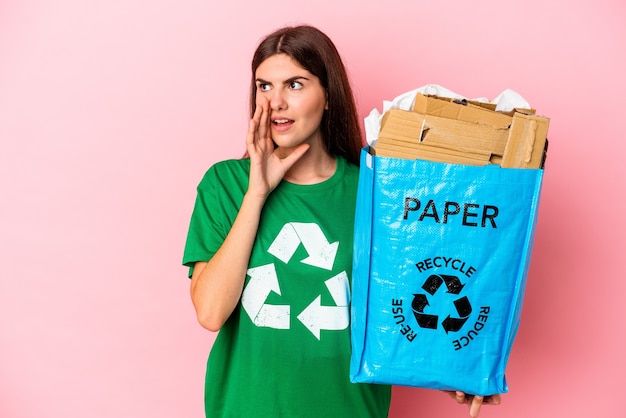 Young caucasian woman recycled cardboard isolated on pink wall is saying a secret hot braking news and looking aside