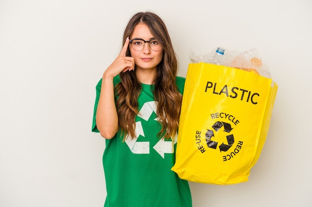 Foto giovane donna caucasica che ricicla un pieno di plastica isolato su sfondo bianco indicando il tempio con il dito, pensando, concentrato su un compito.