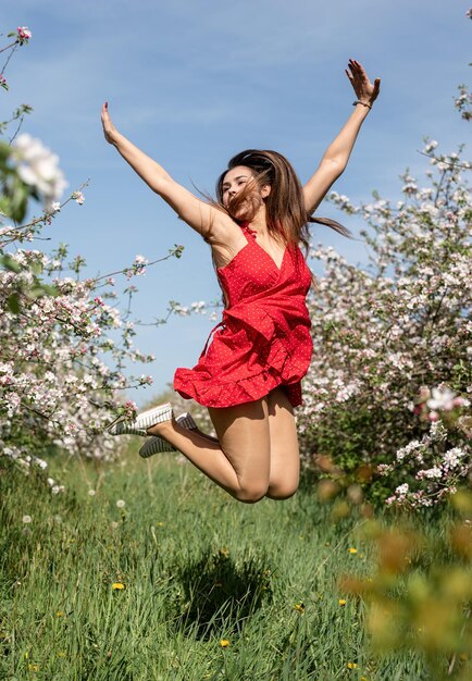 Young caucasian woman in red dress enjoying the flowering of an apple trees jumping