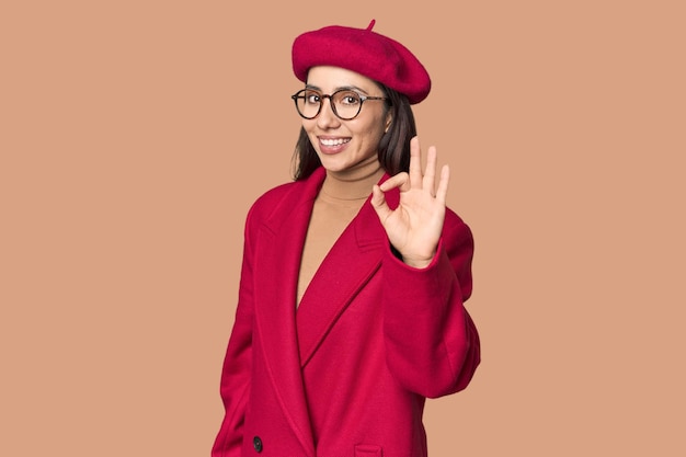 young Caucasian woman in red trench coat and beret cheerful and confident showing ok gesture