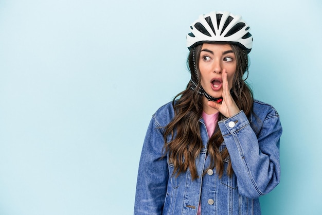 Young caucasian woman rinding a bike isolated on blue\
background is saying a secret hot braking news and looking\
aside