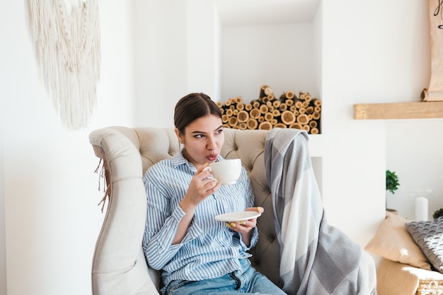 Young caucasian woman sitting on armchair in living room and drinking coffee, enjoying the sunny morning.