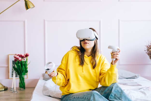 젊은 백인 여자는 침대에 앉아 VR 헤드셋을 사용하여 컨트롤러를 잡고 가상 현실을보고 있습니다.