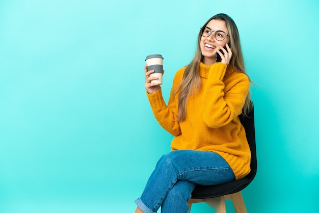 持ち帰り用のコーヒーと携帯電話を保持している青い背景で隔離の椅子に座っている若い白人女性