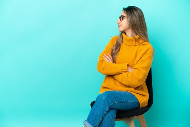 Young caucasian woman sitting on a chair isolated on blue background in lateral position