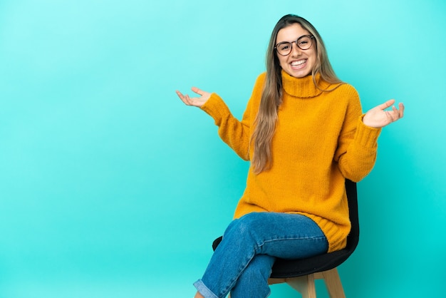 Young caucasian woman sitting on a chair isolated on blue background with shocked facial expression